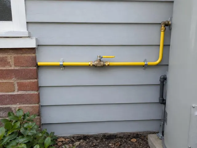 Yellow gas pipe and shutoff valve on grey weatherboard wall beside a brick corner in Rockdale